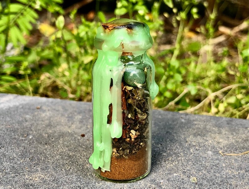 May include: A small glass bottle filled with layers of herbs, spices, and a small green stone. The bottle is sealed with a cork and green wax drippings. The background is blurred green foliage.