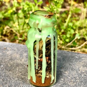 May include: A small glass bottle filled with dried herbs and a brown powder at the bottom. The bottle is sealed with a green wax that has dripped down the sides. The background is blurred green foliage.