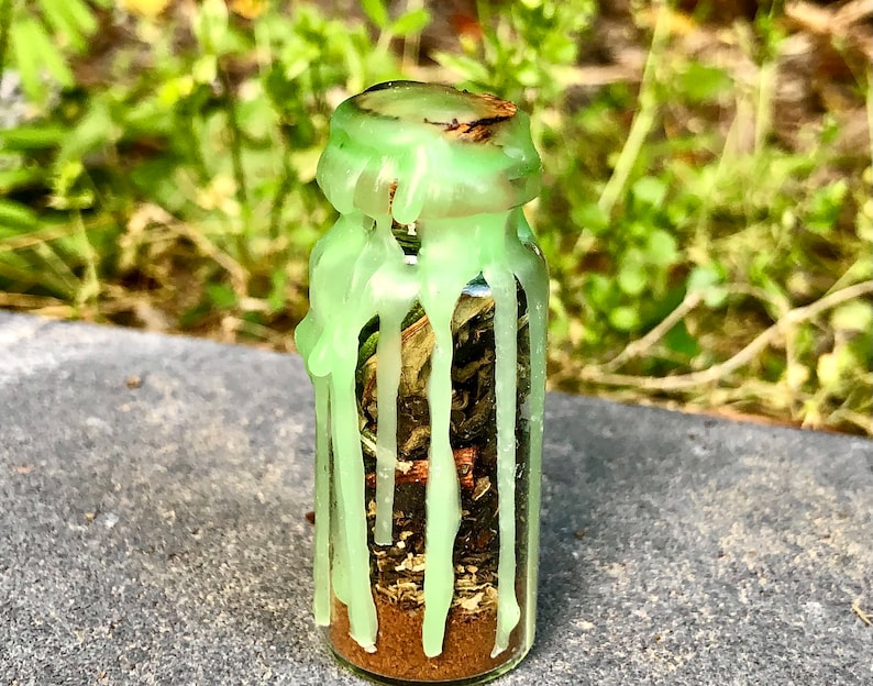 May include: A small glass bottle filled with dried herbs and a brown powder at the bottom. The bottle is sealed with a green wax that has dripped down the sides. The background is blurred green foliage.