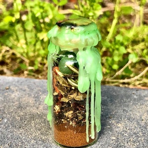 May include: A small glass bottle filled with layers of brown, green, and tan materials, topped with a light green wax seal. The wax is dripped down the sides of the bottle. The bottle contains a green stone and dried herbs.