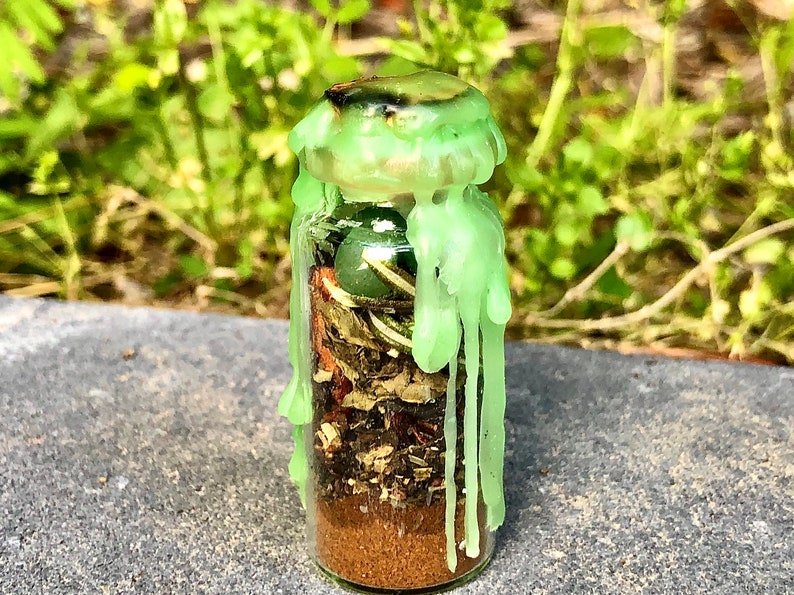 May include: A small glass bottle filled with layers of brown, green, and tan materials, topped with a light green wax seal. The wax is dripped down the sides of the bottle. The bottle contains a green stone and dried herbs.