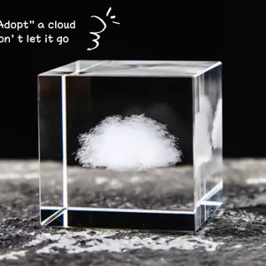May include: A clear glass cube with a 3D image of a white cloud inside. The cube is sitting on a gray textured surface. The text "Adopt" a cloud Don't let it go" is written above the cube.