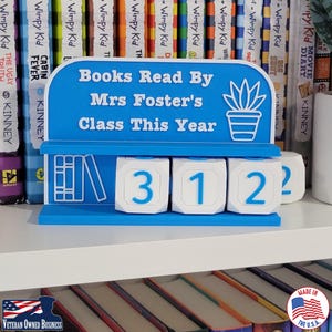 May include: A blue and white 3D-printed sign displays "Books Read By Mrs Foster's Class This Year." It features a plant graphic and rolling number cubes showing "312". The sign is on a shelf with books.