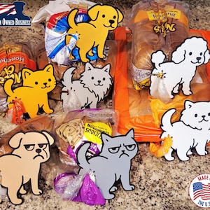 May include: A collection of whimsical wooden pet-shaped bag clips in various colors, including yellow, white, and gray. The clips feature dog and cat designs, with some displaying grumpy expressions. The image also includes loaves of bread.