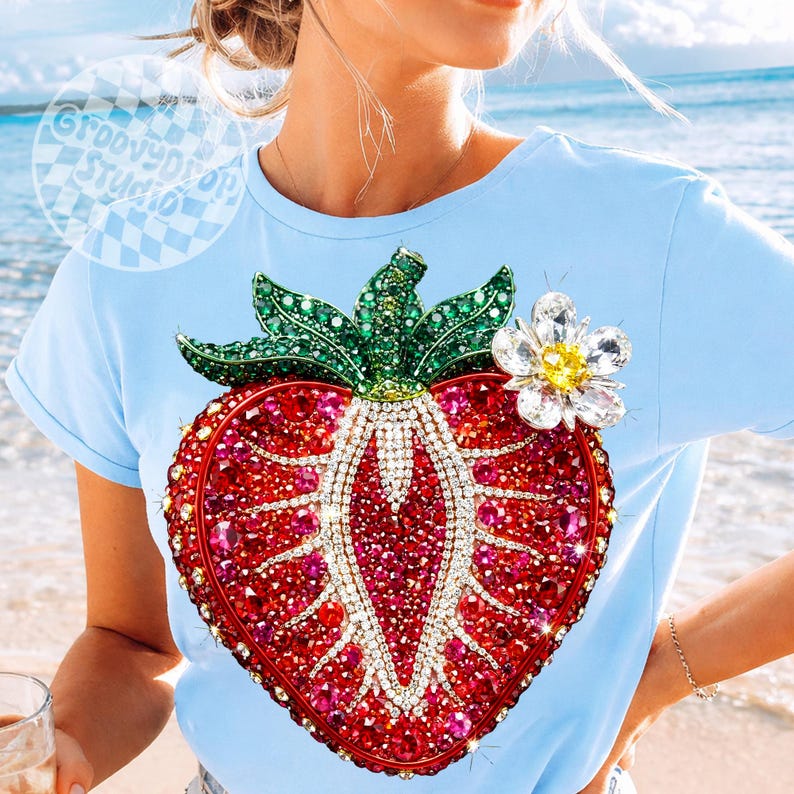 May include: Light blue t-shirt featuring a large, sparkling strawberry design. The strawberry is embellished with red and pink gems, green leaves, and a white flower with a yellow center. The shirt has a crew neck and short sleeves.