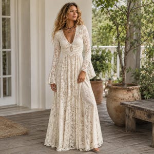 May include: A floor-length cream-colored lace dress featuring long bell sleeves, a deep V-neckline, and a fitted bodice that transitions into a flowing skirt. The intricate floral lace pattern covers the entire garment, which is worn with a long necklace.