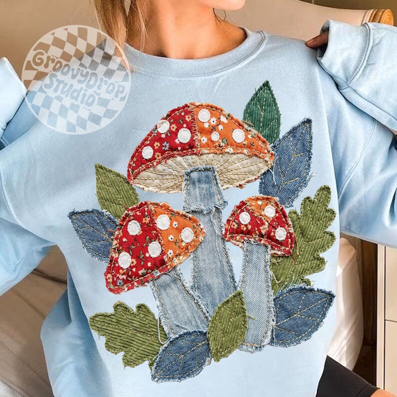 May include: Light blue sweatshirt featuring a whimsical mushroom design. The mushrooms have red and orange caps with white polka dots, denim stems, and are surrounded by green and blue leaves. The text "GroovyDrop Studio" is in a circle on the upper left.