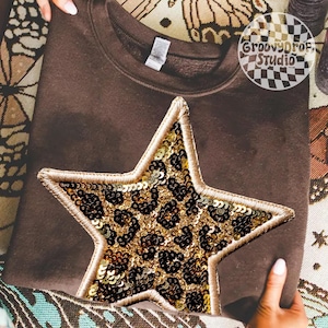 May include: A brown sweatshirt with a large star applique. The star is filled with gold sequins and a leopard print design. The star is outlined with gold thread. The sweatshirt is folded and held by hands.