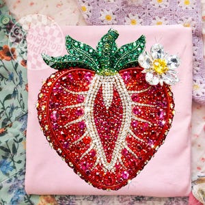 May include: A light pink t-shirt featuring a large, bedazzled strawberry design. The strawberry is made of red and pink rhinestones, with a green rhinestone stem and a white and yellow flower. The design is outlined in red.