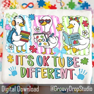 May include: White t-shirt with a colorful graphic featuring three cartoon ducks. One duck wears headphones and holds a pop-it toy, another wears heart-shaped sunglasses, and the third reads a book. The text "IT'S OK TO BE DIFFERENT" is displayed.