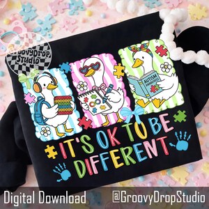 May include: Black sweatshirt with a colorful graphic featuring three cartoon ducks with puzzle pieces, headphones, and books. The text "IT'S OK TO BE DIFFERENT" is displayed in vibrant colors. The design includes the words "Autism Awareness".
