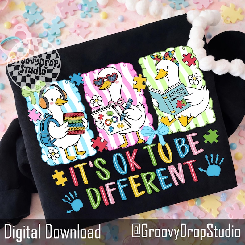 May include: Black sweatshirt with a colorful graphic featuring three cartoon ducks with puzzle pieces, headphones, and books. The text "IT'S OK TO BE DIFFERENT" is displayed in vibrant colors. The design includes the words "Autism Awareness".