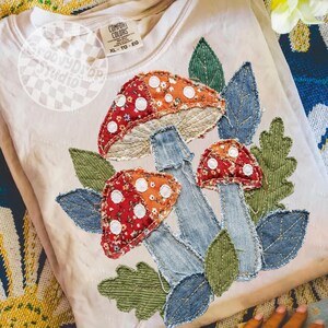 May include: A beige Comfort Colors sweatshirt featuring an appliqu&eacute; design of three mushrooms with red and orange caps, white spots, and denim stems. The design includes green and denim leaves. The sweatshirt has a tag that reads XL-TG-EG.