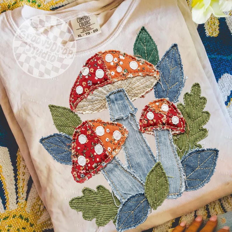 May include: A beige Comfort Colors sweatshirt featuring an appliqu&eacute; design of three mushrooms with red and orange caps, white spots, and denim stems. The design includes green and denim leaves. The sweatshirt has a tag that reads XL-TG-EG.