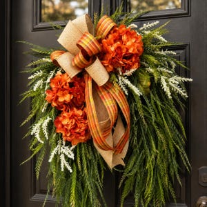 Peut inclure: Une couronne de porte en forme de larme composée de fleurs d'hortensia orange vif, de long feuillage vert semblable à du pin et de brins plumeux blancs. Un grand nœud décoratif en toile de jute beige et ruban à carreaux orange, jaune et vert.
