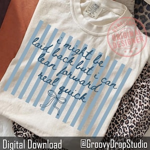 以下が含まれることがあります： クリーム色のTシャツ。水色の縦縞模様と手書きの文字「i might be laid back but i can lean forward real quick」がプリントされています。リボンのグラフィックが下にあります。Tシャツは折りたたまれています。