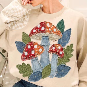 May include: Beige sweatshirt with a colorful mushroom design. The mushrooms have red and orange caps with white polka dots, denim stems, and are surrounded by green and blue leaves. The text "Groovy Drop Studio" is in the upper left corner.