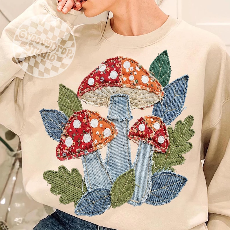 May include: Beige sweatshirt with a colorful mushroom design. The mushrooms have red and orange caps with white polka dots, denim stems, and are surrounded by green and blue leaves. The text "Groovy Drop Studio" is in the upper left corner.