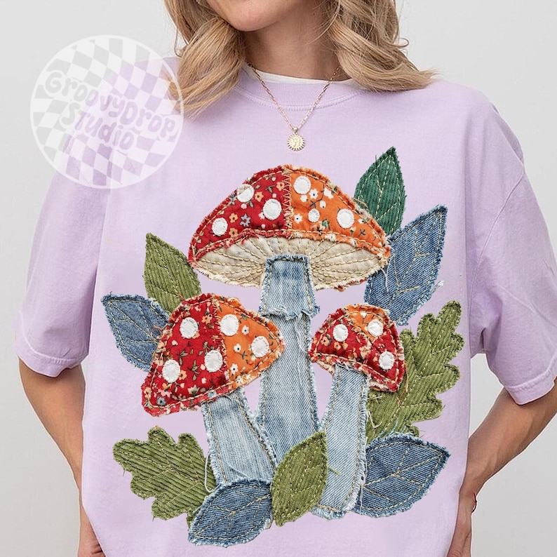 May include: A light purple t-shirt featuring a whimsical mushroom design. The mushrooms have red and orange caps with white polka dots, denim stems, and are surrounded by green leaves. The shirt has the text "Groovy Drop Studio".