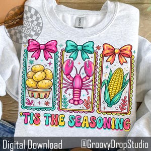 May include: Pale grey sweatshirt with a vibrant graphic of potatoes, a lobster, and corn, each adorned with a bow. The phrase "'Tis the Seasoning" is displayed below. Digital download.