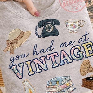 May include: Gray sweatshirt with embroidered vintage-themed patches, including a rotary phone, teacup, hat, books, gramophone, and a lamp. The phrase "you had me at VINTAGE" is embroidered in blue and floral-patterned letters.