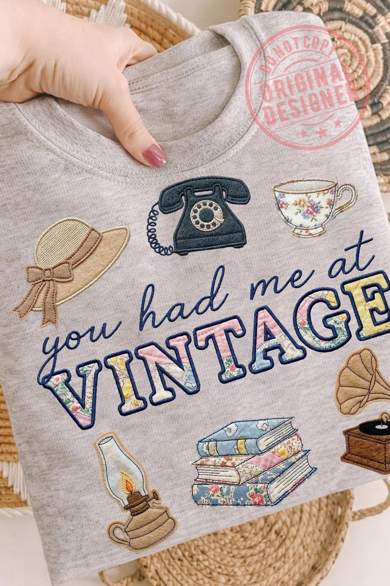 May include: Gray sweatshirt with embroidered vintage-themed patches, including a rotary phone, teacup, hat, books, gramophone, and a lamp. The phrase "you had me at VINTAGE" is embroidered in blue and floral-patterned letters.