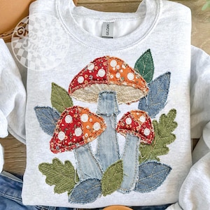 May include: A light gray sweatshirt featuring an appliqu&eacute; design of three mushrooms and leaves. The mushroom caps are red and orange with white polka dots. The stems are denim, and the leaves are green and blue.