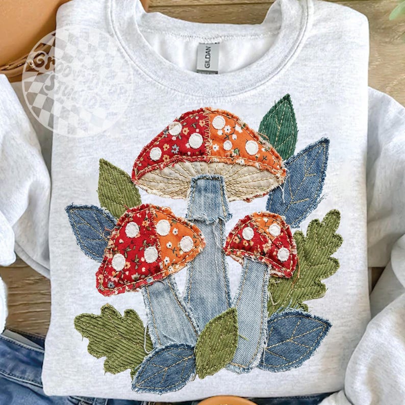 May include: A light gray sweatshirt featuring an appliqu&eacute; design of three mushrooms and leaves. The mushroom caps are red and orange with white polka dots. The stems are denim, and the leaves are green and blue.