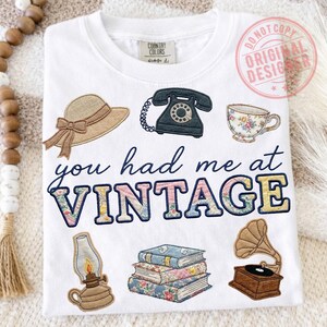 May include: White t-shirt with embroidered vintage-themed designs, including a hat, telephone, teacup, lamp, books, and a gramophone. The phrase "you had me at VINTAGE" is embroidered in a cursive font. A "Country Colors" tag is visible.