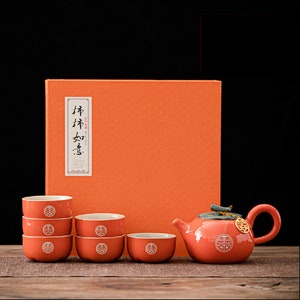 Peut inclure: Un ensemble de tasses à thé et d'une théière orange avec un caractère chinois pour "double bonheur" sur chaque pièce. L'ensemble est emballé dans une boîte orange avec le même caractère sur le devant.
