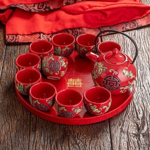May include: A red and gold Chinese tea set with a teapot and 10 cups. The cups and teapot have intricate designs and the word "囍" (double happiness) is on the tray.
