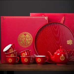 Puede incluir: Un juego de té rojo con tetera, tazas y bandeja. El juego está decorado con caracteres chinos dorados, incluido el símbolo de la doble felicidad. El juego se presenta en una caja roja.