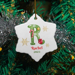 May include: White ceramic snowflake ornament with a green letter "R" decorated with candy canes and a red boot. The ornament also includes the name "Rachel" and the year "2023".