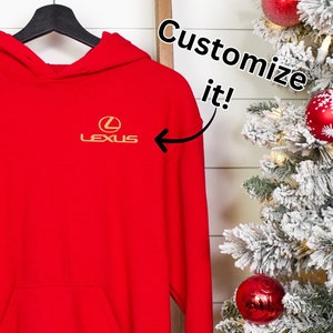 May include: Red hooded sweatshirt with a gold Lexus logo on the front. The text "Customize it!" is written in a white circle with a black outline. The sweatshirt is hanging on a hanger in front of a white wall with a Christmas tree on the right.