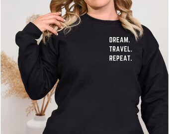 Könnte beinhalten: Schwarzes Langarm-Sweatshirt mit den Wörtern "DREAM. TRAVEL. REPEAT." in Weiß aufgedruckt. Das Sweatshirt ist einfarbig schwarz und hat einen Rundhalsausschnitt. Der Text ist vertikal auf der oberen Brust ausgerichtet.