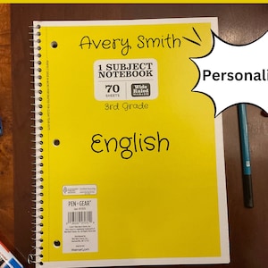 May include: A yellow 1 subject notebook with 70 wide ruled sheets for 3rd grade English. The notebook has a personalized name Avery Smith on the front. The notebook is on a wooden table with pens and markers.