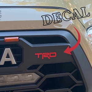 May include: A tan vehicle front grille featuring a matte black honeycomb pattern and a prominent red TRD decal. The grille displays the silver letters TOYOTA, a small orange light, and a red arrow pointing toward the red TRD lettering.