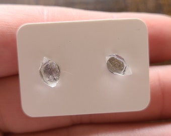Puede incluir: Un par de pequeños pendientes de botón transparentes sobre una tarjeta blanca. Los pendientes se sujetan con plástico transparente. Los pendientes son redondos y tienen un respaldo plateado. La tarjeta se sostiene en una mano.