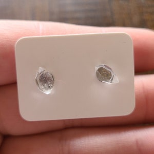 Könnte beinhalten: Ein Paar kleiner, klarer Ohrstecker auf einer weißen Karte. Die Ohrringe werden mit klarem Kunststoff fixiert. Die Ohrringe sind rund und haben eine silberne Rückseite. Die Karte wird in einer Hand gehalten.