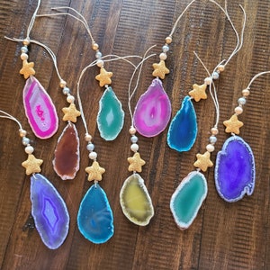 May include: Ten agate slice ornaments with gold star charms and string for hanging. The agate slices are in various colors, including pink, purple, blue, green, and yellow.