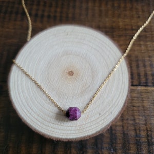 May include: A delicate gold-colored chain necklace featuring a faceted, deep pink gemstone pendant. The necklace is displayed on a small, round wooden slice with a natural wood grain pattern. The pendant is a geometric shape.