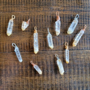 May include: A collection of ten clear quartz crystal pendants wrapped in copper wire. The crystals are various sizes and shapes, and each one has a different copper wire design.