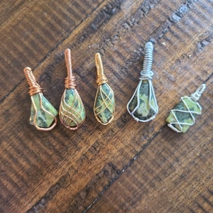 May include: Five green and gold wire wrapped pendants with a variety of stone shapes. The pendants are on a wooden surface.