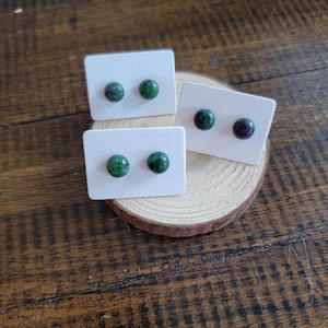 Puede incluir: Tres pares de pendientes de botón con piedras preciosas moteadas verdes y rojas. Cada par está montado en una tarjeta blanca, exhibido sobre una superficie de madera. Los pendientes son redondos y parecen ser de piedra natural.