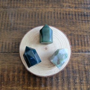 May include: Three polished gemstone towers in shades of green, black, and white are arranged on a small, round wooden slice. The gemstones have faceted surfaces and are displayed against a natural wood background.