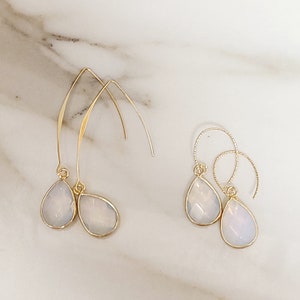 May include: Two pairs of gold-toned earrings with pear-shaped, opalescent pendants. One pair features long, curved hooks, while the other has smaller, circular hoops. The earrings are displayed on a white marble surface.