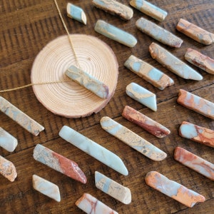 May include: A collection of polished, light blue and brown, natural stone pendants. The stones have a marbled pattern and are arranged on a wooden surface. One pendant is on a gold chain necklace.
