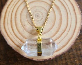 Puede incluir: Un collar de cadena dorada con un colgante de cristal de cuarzo transparente. El colgante es una punta doblemente terminada con una banda dorada en el medio.