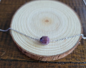 May include: A silver chain necklace with a faceted, dark purple gemstone pendant. The necklace is displayed on a wooden slice with a natural wood grain pattern. The background is a dark wood surface.