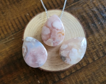 May include: Three teardrop-shaped pendants made of pink and white agate. The pendants are on a wooden surface.